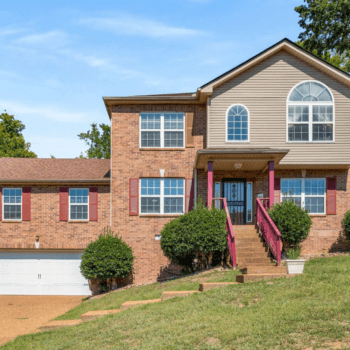 Front view of a two-story brick home with red shutters and a charming front porch, featuring a well-maintained lawn and a two-car garage, located in a quiet cul-de-sac. Listed by Patricia Omishakin